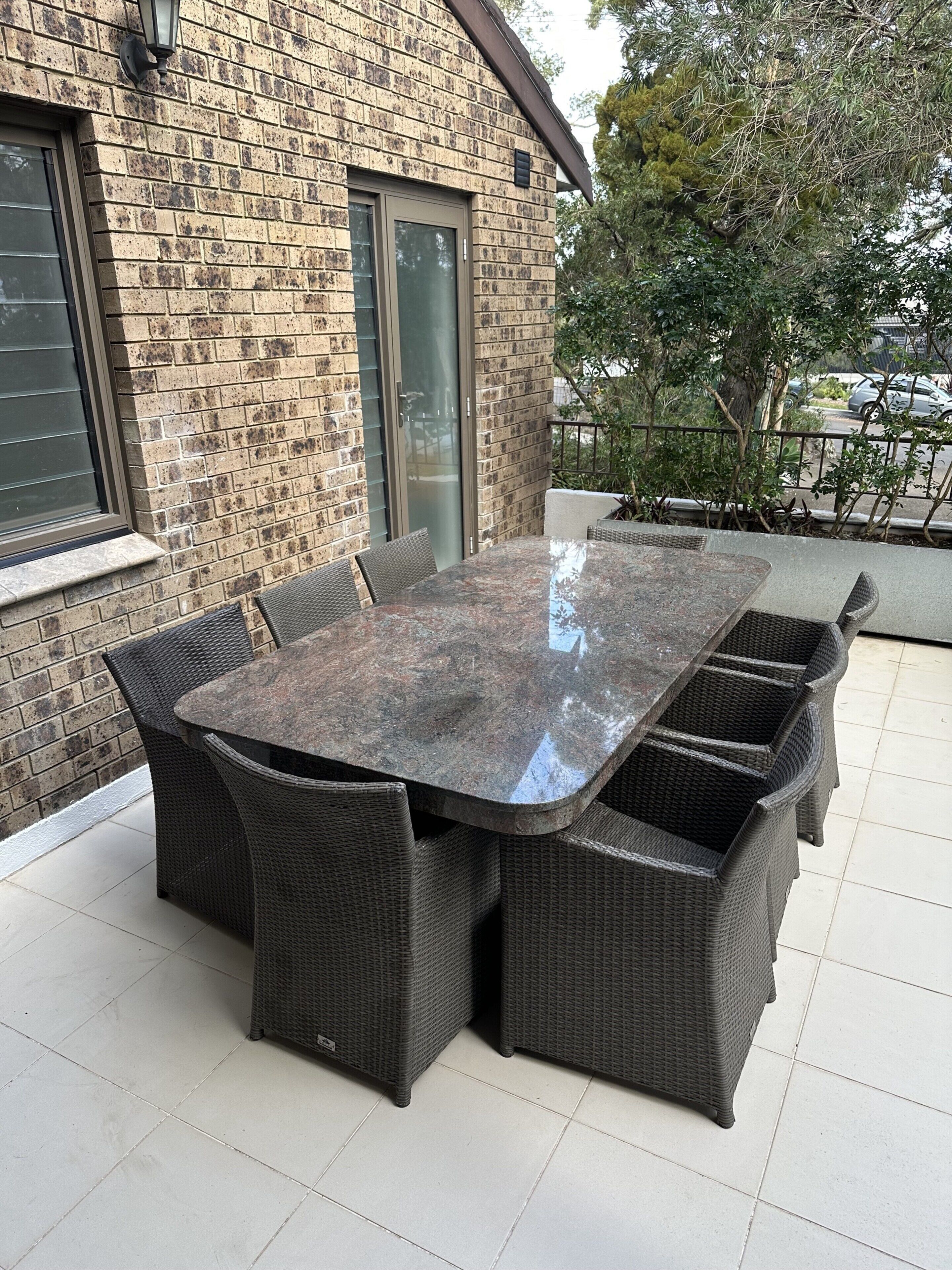 Un patio con una mesa grande y oscura, de forma rectangular, rodeada de sillas de mimbre.
