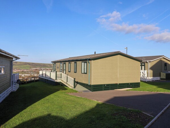 A single-story mobile home with a green roof and beige walls, a small porch with a white railing, and a view of a distant town.