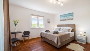 Un dormitorio con una cama, una silla, una mesita, una mesita de noche, un cuadro y una ventana.