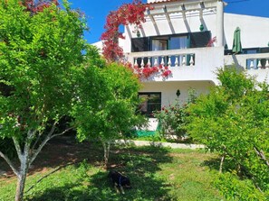 A two-story building with a balcony, surrounded by greenery and a dog in the yard.