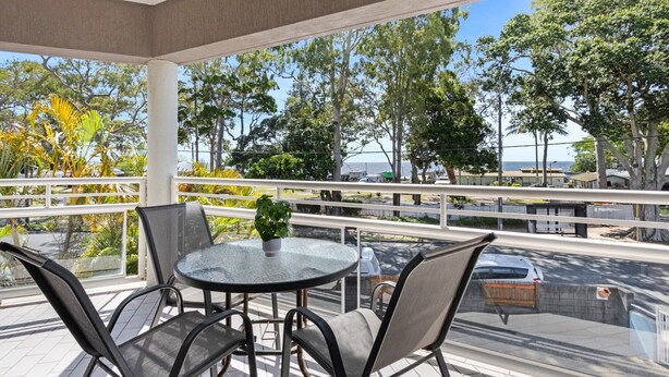 A covered outdoor seating area with a round table and chairs, overlooking a street with trees and parked cars. - Beachside 2-bed Apartment With Secure Parking (Hervey Bay)