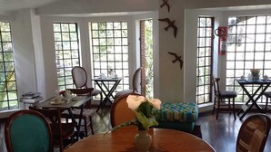 Un coin repas avec une table ronde en bois, des chaises et un luminaire suspendu. On y trouve des fenêtres avec des vitres à barreaux et un extincteur fixé au mur.