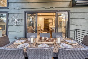 An outdoor dining area with wicker furniture, a central table with candles and pine cones, and a view into the house through glass doors. - Luxury Mountain Lodge | Views, Hot Tub & Firepit (Blue Ridge)