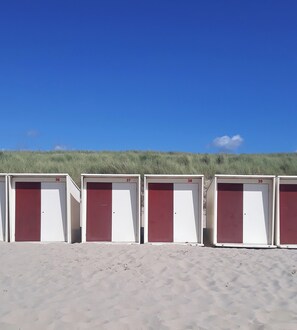 Fila de cabañas de playa numeradas con puertas blancas y rojas, sobre un cielo azul despejado y una duna cubierta de césped al fondo.