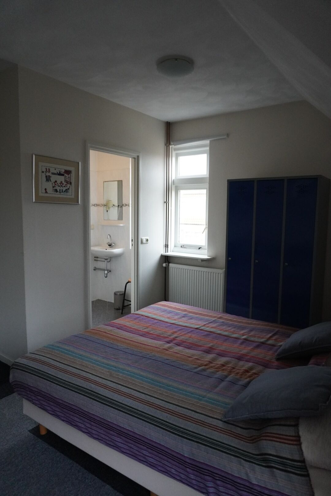 Ein Schlafzimmer mit Bett, Fenster, Heizkörper, Kleiderschrank und angrenzendem Badezimmer.
