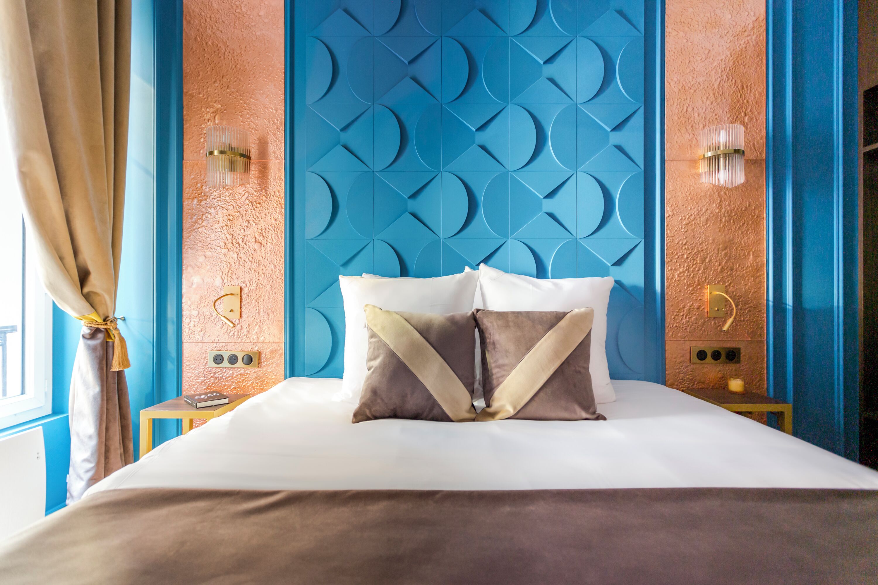 A neatly made bed with two pillows, a small bedside table with a book, and a wall with a geometric pattern.
