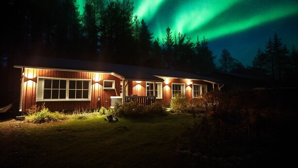 Een houten hutje met een groot raam, verlicht door buitenverlichting, omgeven door een bos en onder een nachthemel met groene poollicht.