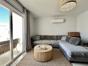 A modern living room with a grey sectional sofa, a round wooden coffee table, and a woven ceiling light fixture. - 41 KAP - Stay Paradise Apartment (Paralimni)