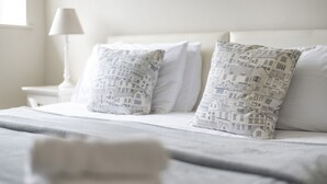 Ein ordentlich bezogenes Bett mit weißer Bettwäsche und gemusterten Kissen, eine Nachttischlampe und ein Fenster im Hintergrund.