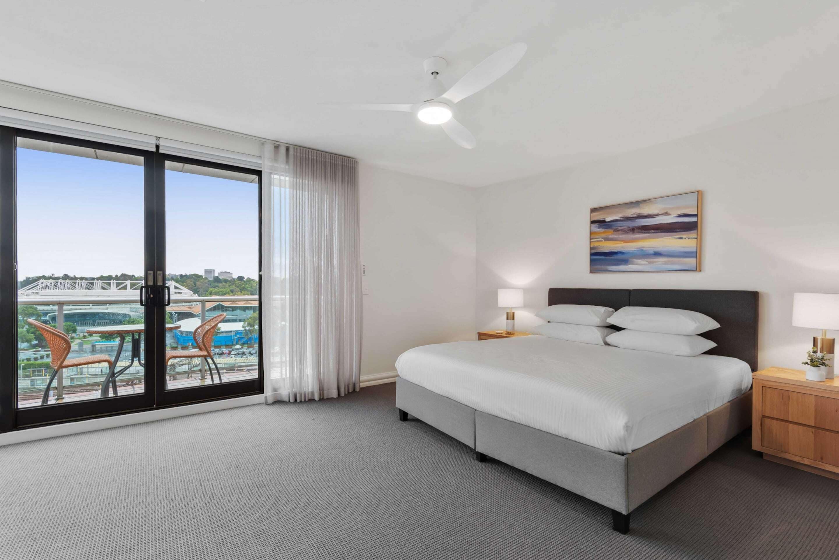 Habitación de hotel con cama, ventilador de techo, vista a un paisaje urbano y balcón con sillas.
