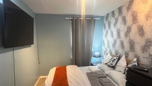 A bedroom with a bed, a flat-screen TV mounted on the wall, a nightstand with a lamp, and a dresser.
