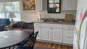 A kitchen with white cabinets, a granite countertop, a sink, and a refrigerator. There is a dining table with chairs and a lamp on a side table.