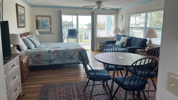 A room with a bed, a table with chairs, a sofa, a ceiling fan, and a view through a window.