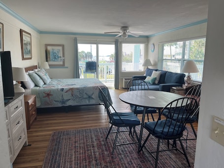 A room with a bed, a table with chairs, a sofa, a ceiling fan, and a view through a window.