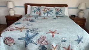 A bed with a starfish and coral themed quilt, wooden headboard, two bedside tables with lamps, and a framed picture on the wall.