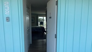 A hallway with a wooden floor leading to a room with a bed and a chair. A door with a number sign is visible on the left.