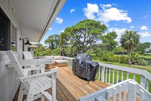 Una terraza de madera con mobiliario exterior, una parrilla y vistas a un gran árbol y al paisaje.