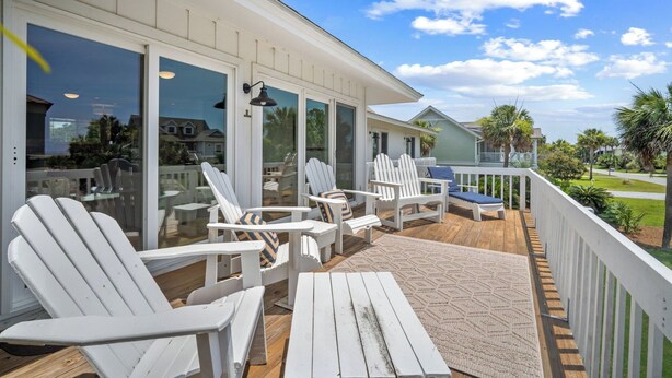 En overdækket terrasse med hvide Adirondack-stole, et træbord og en glasdør, der fører til en patio.