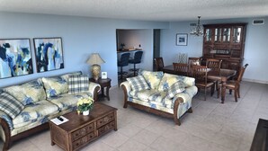 Un salon avec deux canapés, une table basse et une table à manger avec des chaises. Les murs sont ornés de tableaux abstraits et un lustre illumine la pièce.