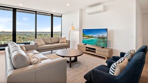 Een moderne woonkamer met een grote televisie, comfortabele zitplaatsen en een stadsgezicht door grote ramen.