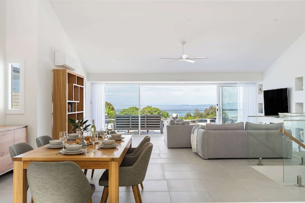 A modern living room with a dining area, a sofa, a TV, and a view of the outdoors.