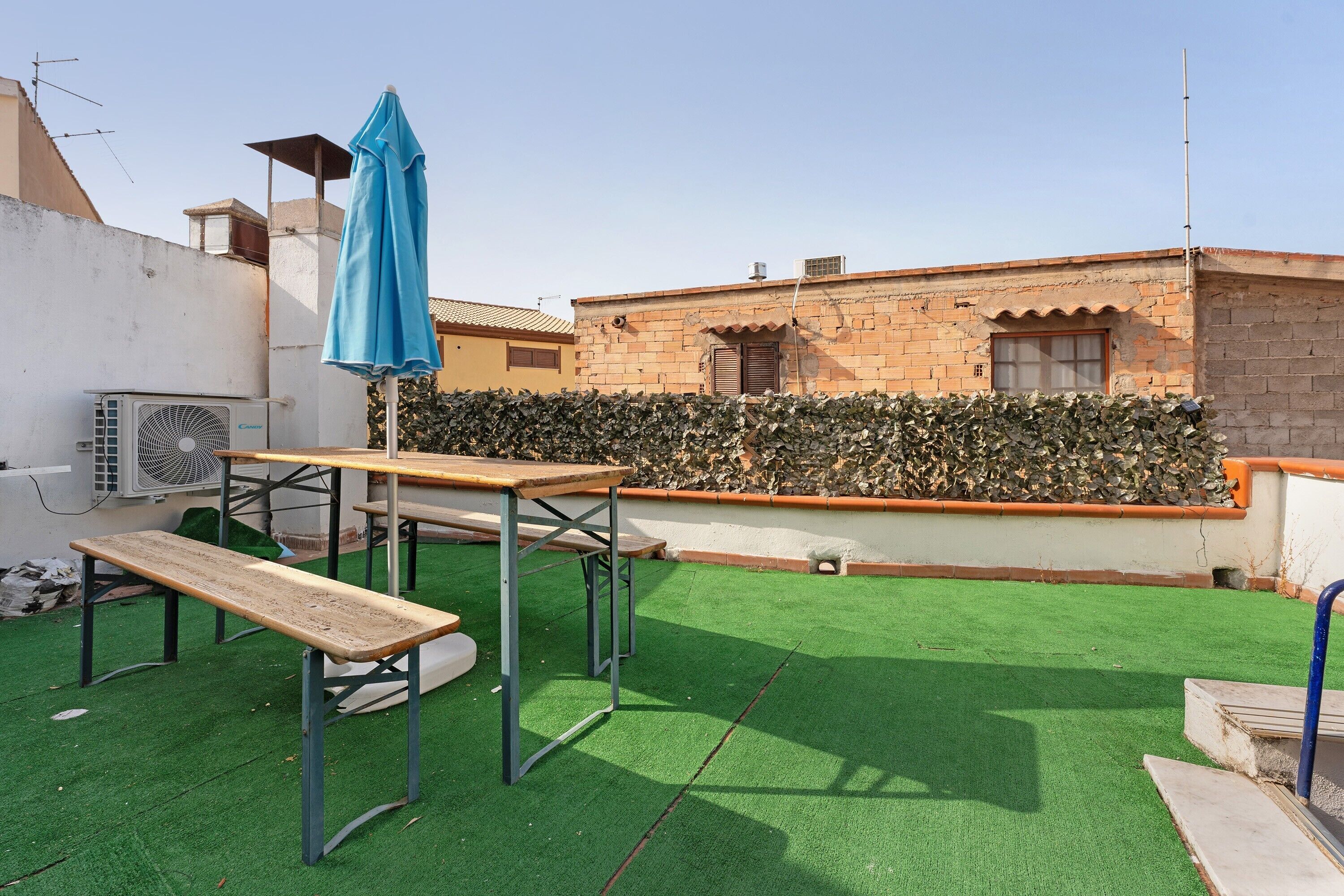 Terraza en la azotea con césped artificial, un banco de madera, una mesa y un paraguas azul.