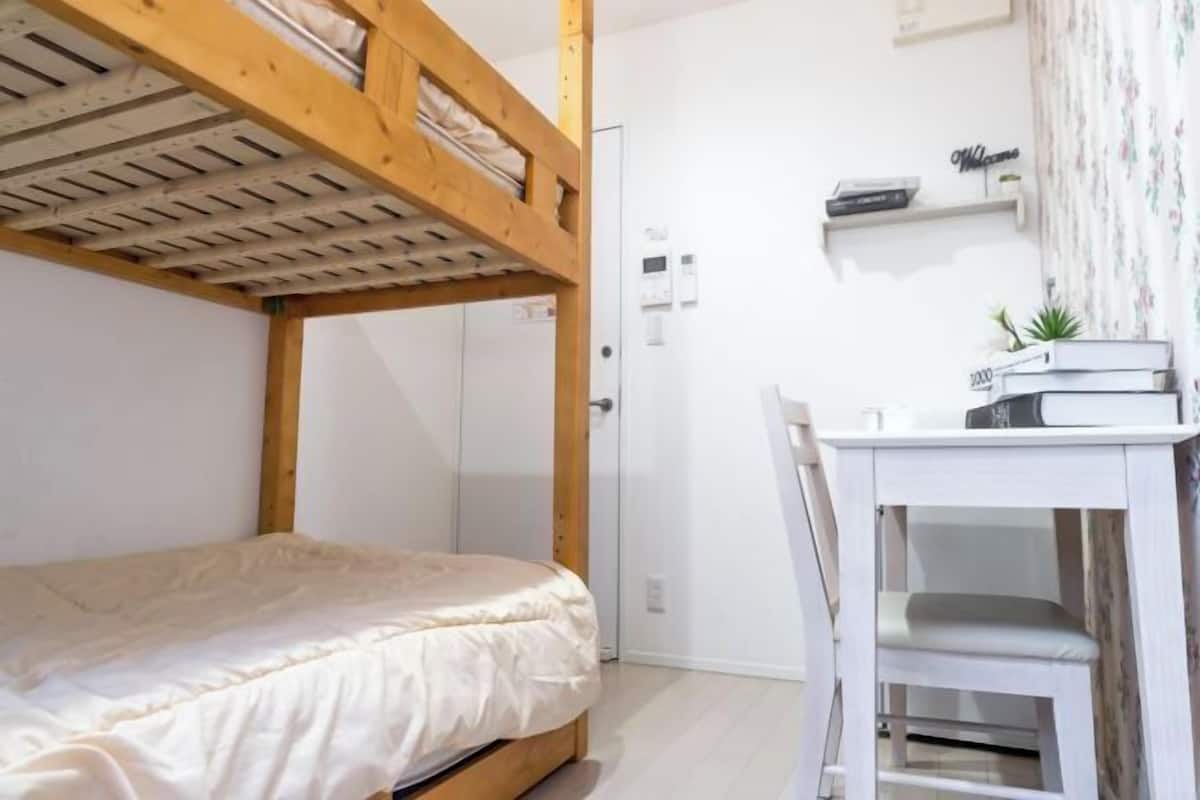 A bunk bed with a wooden headboard, a white desk with a chair, and a small shelf with books and a plant.