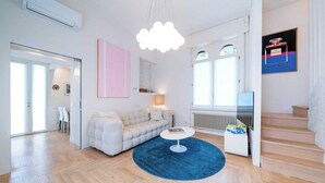 Una sala moderna con un sofá, una mesa de centro y una alfombra azul. Se observa una escalera a la derecha y una puerta que da a otra habitación.