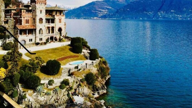 Un edificio grande e histórico con una torre, ubicado junto a un lago y montañas al fondo.
