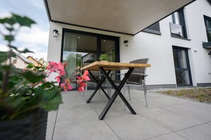 A modern outdoor patio with a wooden table and chairs, a potted plant, and a building with large windows and a door.