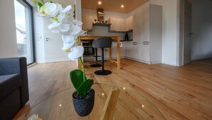 Un salon moderne avec un plancher en bois, une table basse en verre, une orchidée en pot, un canapé gris et un coin cuisine à l’arrière-plan.
