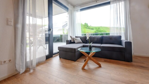 Un salon moderne avec un canapé d’angle gris, une table basse en verre et une vue sur un paysage verdoyant grâce à de grandes fenêtres.