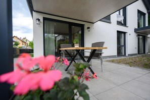 An outdoor patio with a table and chairs, a glass door leading to a building, and a garden area with pink flowers.