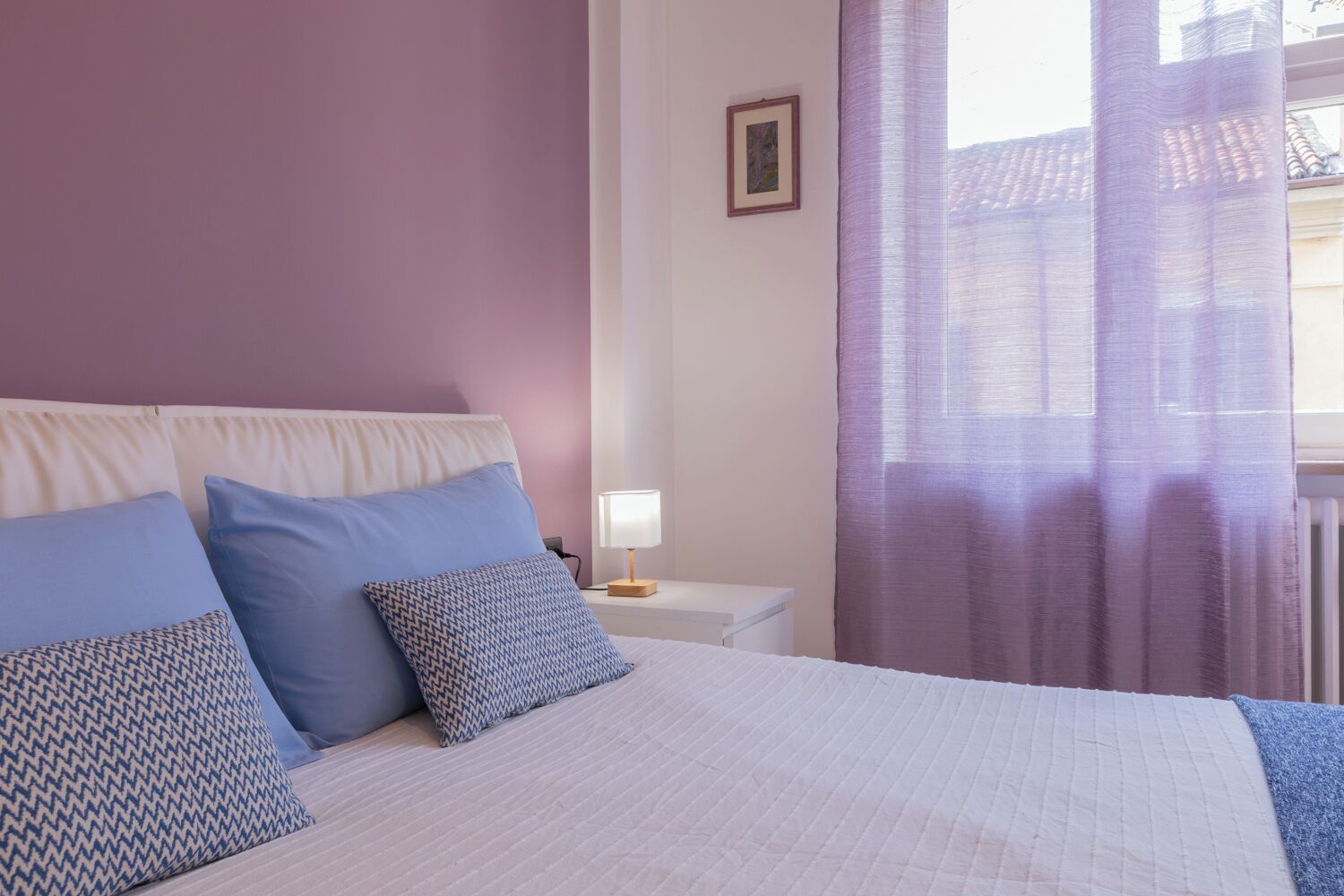 A bedroom with a bed, pillows, a bedside table with a lamp, and a window with curtains.