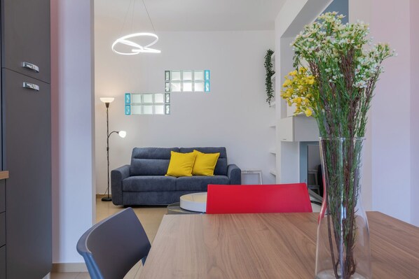 A modern living room with a grey sofa, yellow cushions, a wooden dining table, and a vase with flowers.