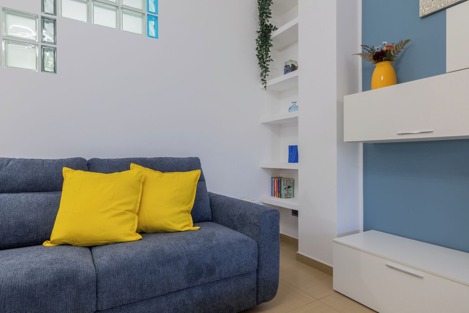 A modern living room with a grey sofa, two yellow cushions, a white bookshelf, and a blue accent wall.