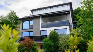 Una casa moderna de dos pisos con balcón, rodeada de exuberante vegetación.