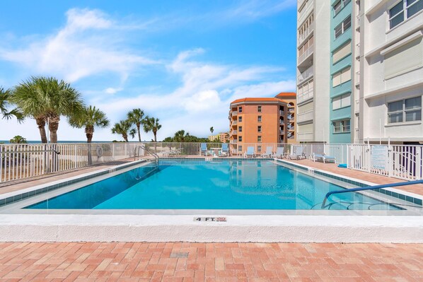 A swimming pool with a white deck, surrounded by palm trees and a building with balconies.