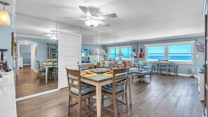 A spacious living area with a dining table, chairs, a ceiling fan, and a view of the ocean through large windows. - Treasure Sands #502 (Treasure Island)