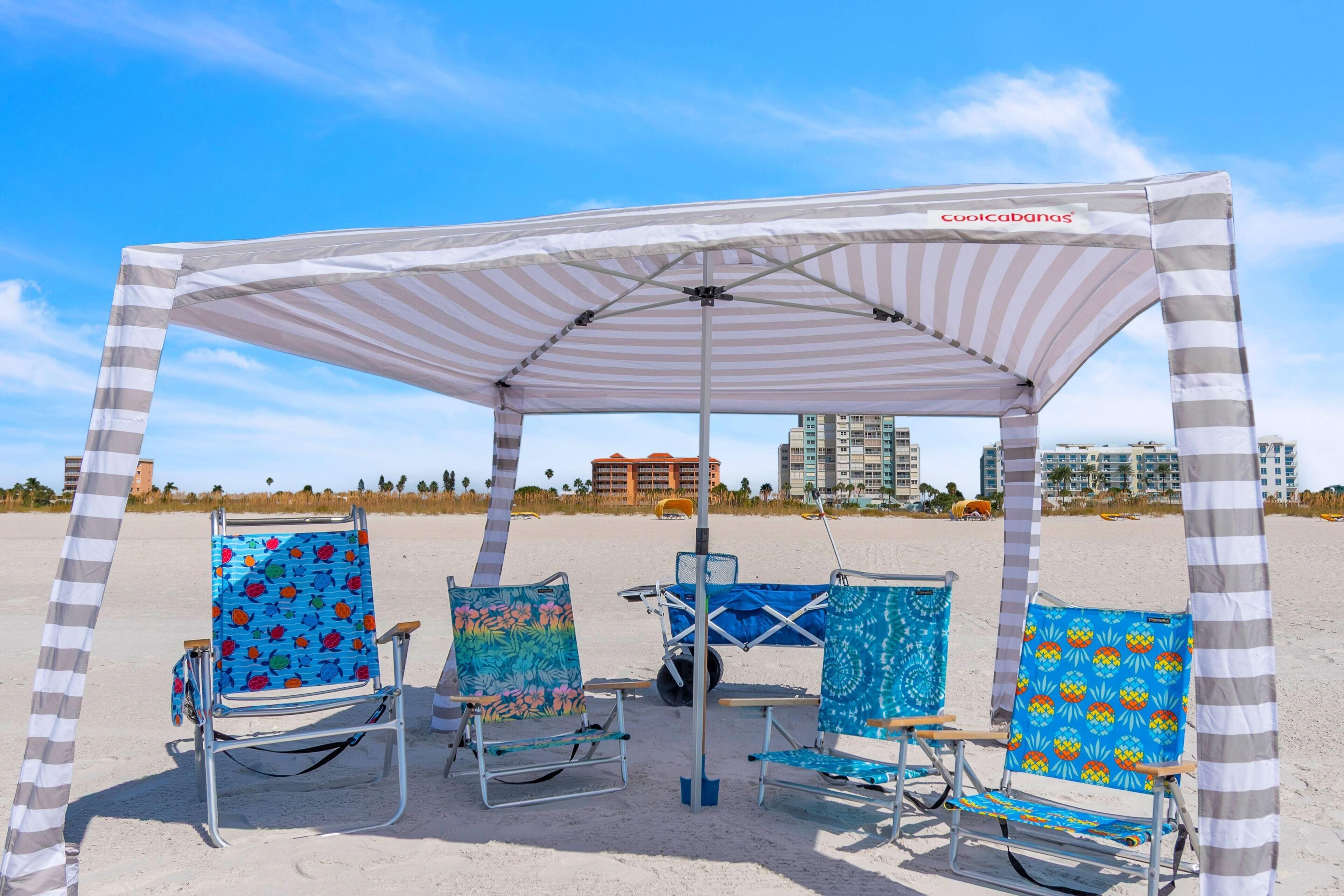 A beach umbrella with a striped canopy, three beach chairs with colorful patterns, and a clear view of the beach and buildings in the background.