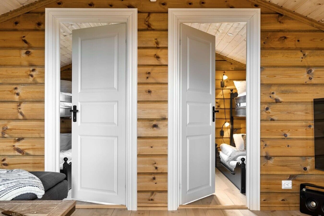 A wooden cabin interior with two white doors, a bed, a table, and a lamp.