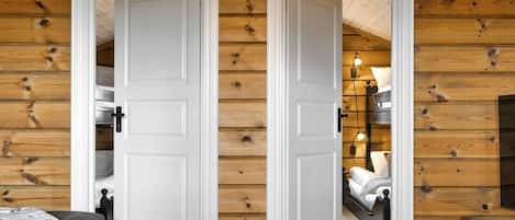 A wooden cabin interior with two white doors, a bed, a table, and a lamp.