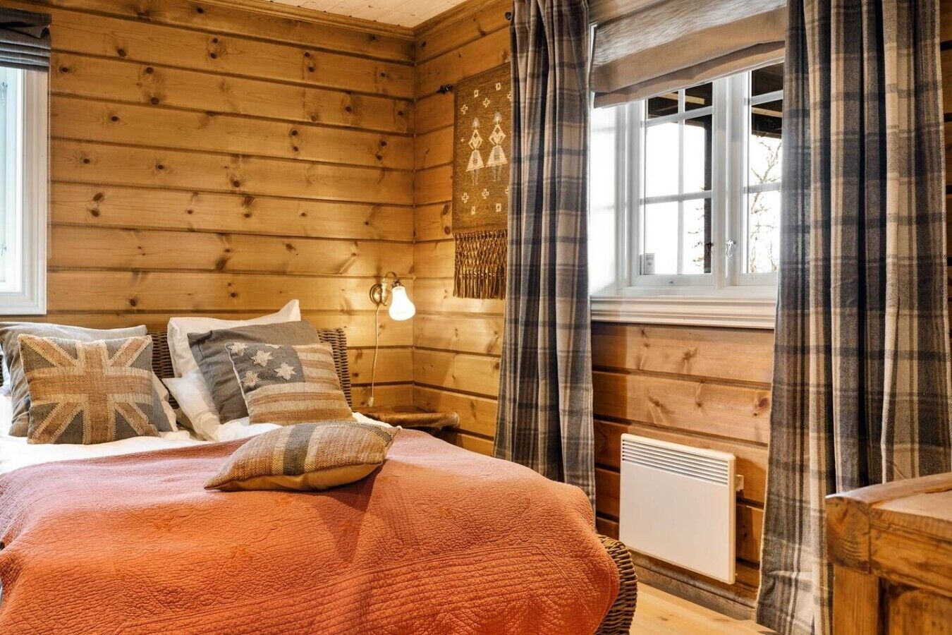 A wooden cabin bedroom with a bed, pillows, a wall-mounted light, and a window with curtains.