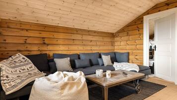 A cozy cabin interior with a wooden ceiling, a sofa with cushions, a coffee table, and a blanket.