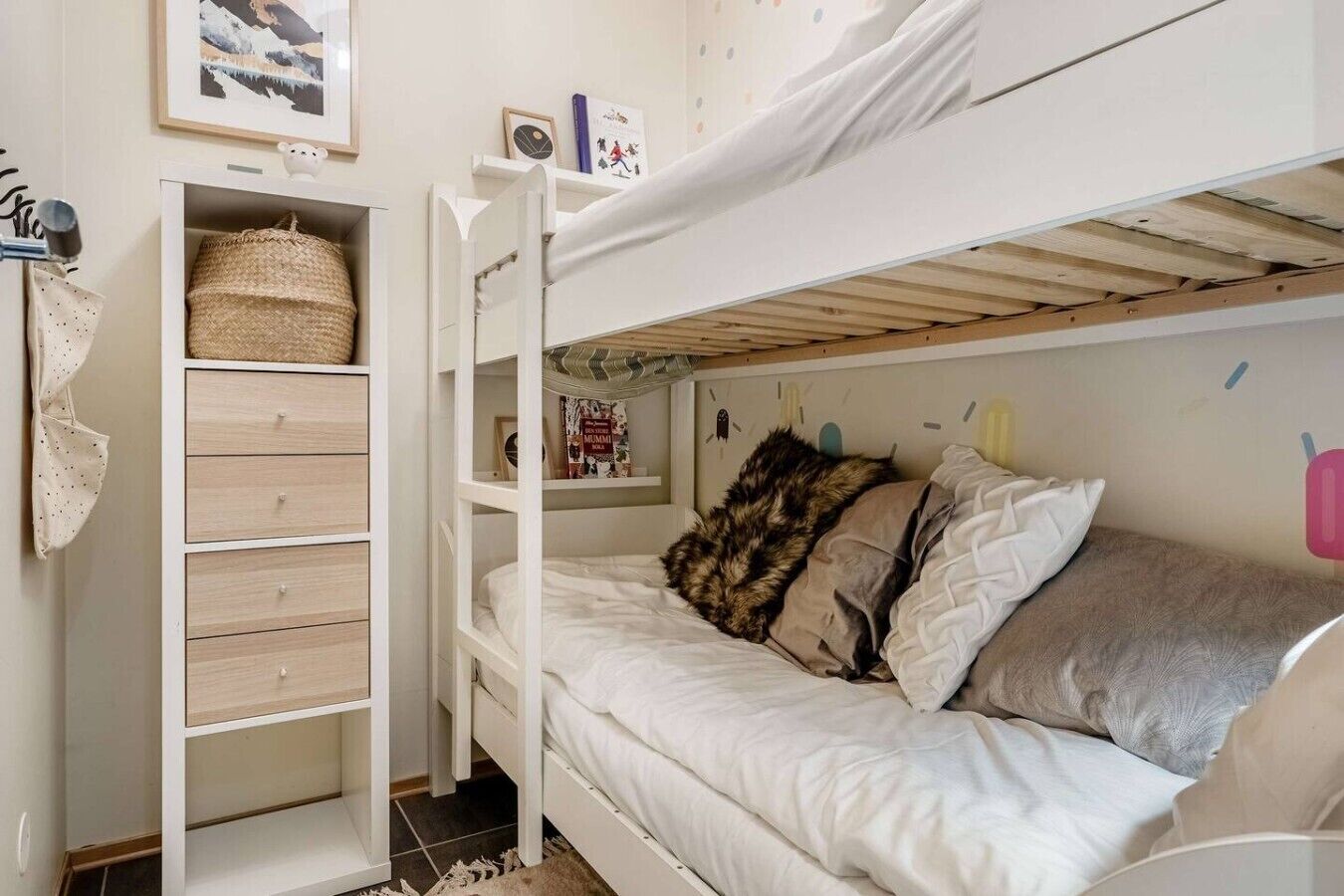 Una cama litera con un módulo de almacenamiento a un lado, un estante en la pared con un libro y un pequeño adorno.
