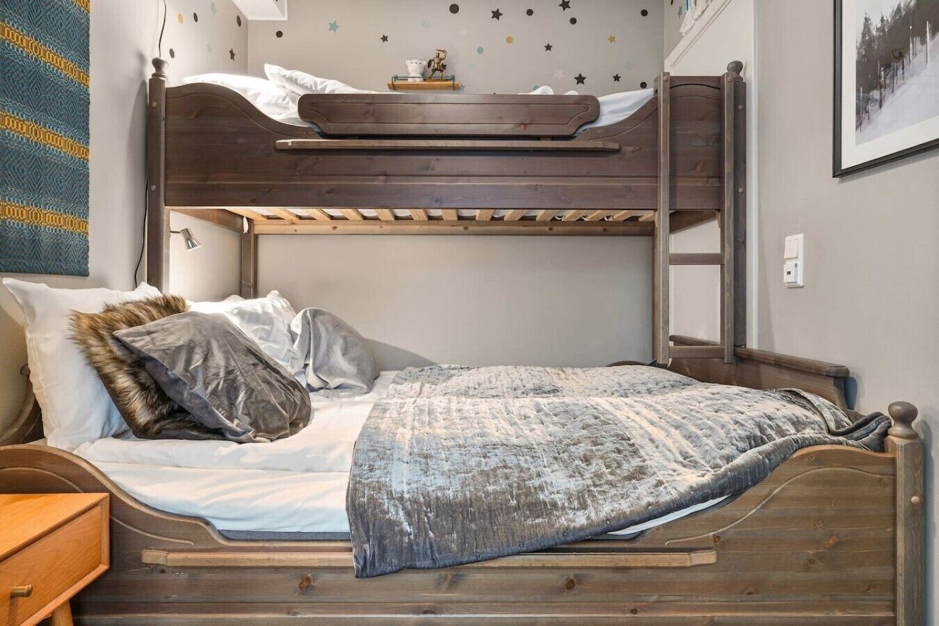 Ein Stockbett mit Holzbettrahmen, ein unteres Bett mit gemustertem Überwurf, Kissen und ein Nachttisch mit Schublade.