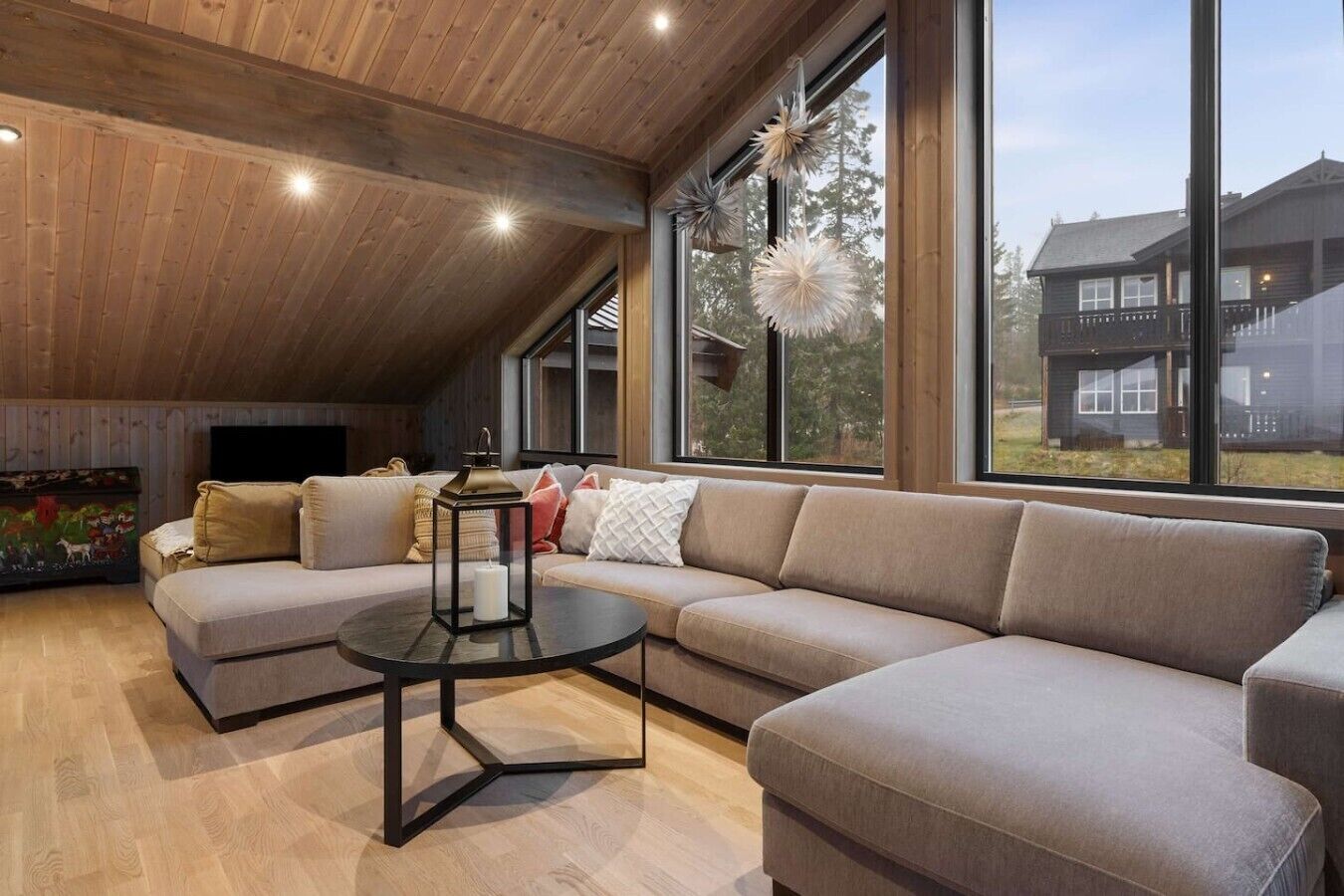 A modern living room with a sectional sofa, a round coffee table, and large windows offering a view of a house.