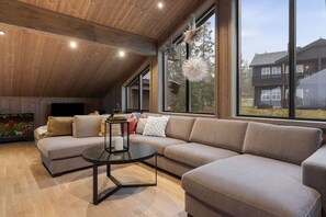 A modern living room with a sectional sofa, a round coffee table, and large windows offering a view of a house.