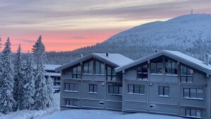 Rakennus, jossa on tasakatto ja useita ikkunoita, ja jonka ympärillä on lumipeitteisiä puita ja kumpuilevaa maastoa.