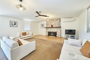 A spacious living room with a stone fireplace, two white sofas, a ceiling fan, and a flat-screen TV.